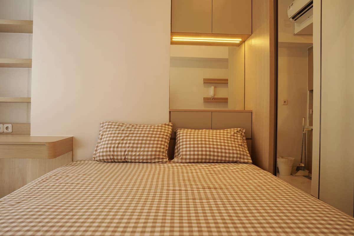 A spacious bed is dressed in checkered bedding, positioned against a light-colored wall. Nearby, shelving and a softly lit area provide functional space. The room showcases a clean and organized design, with warm lighting accentuating the serene atmosphere.