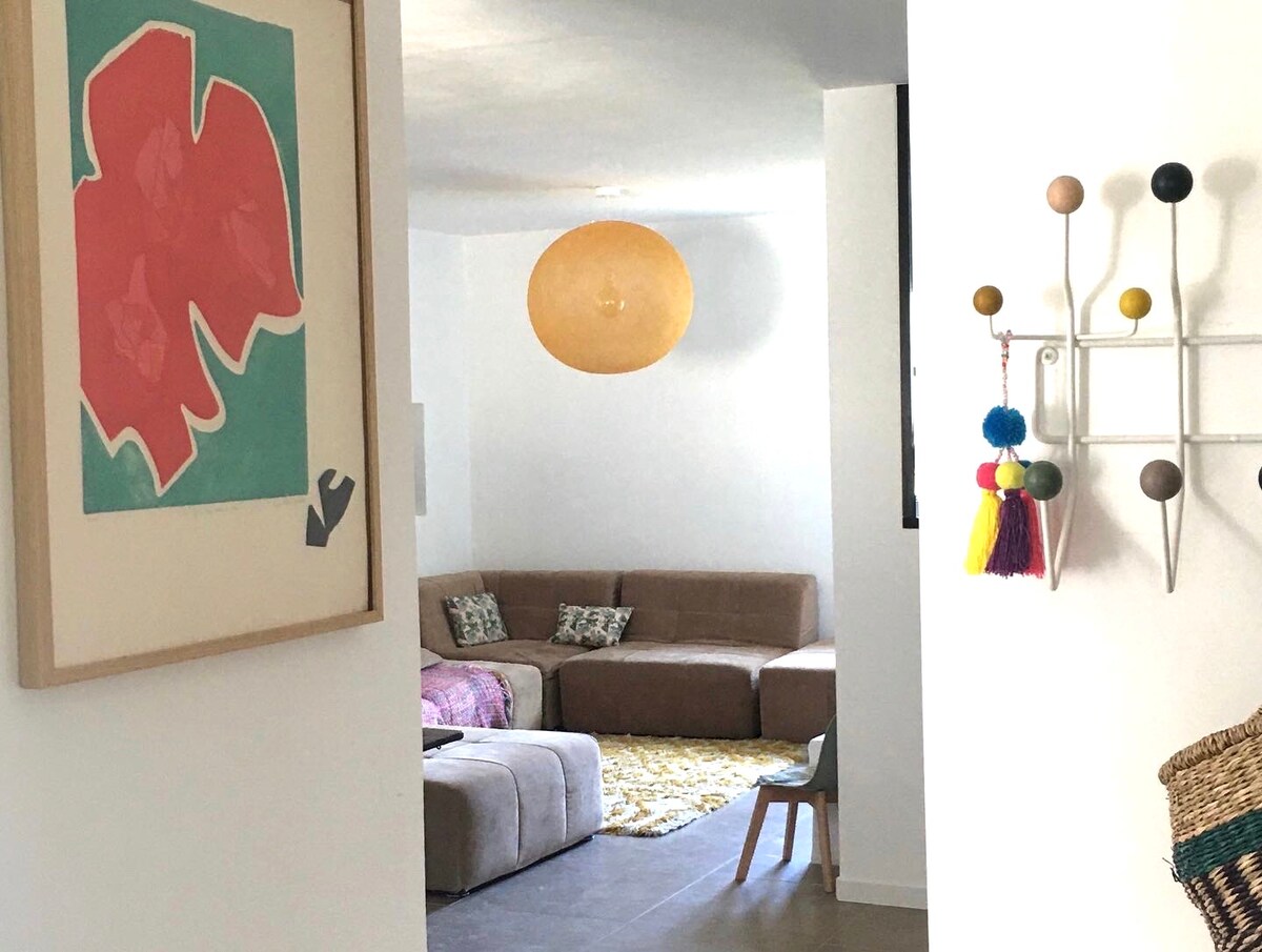 A bright living area is visible, featuring a large sectional sofa complemented by decorative cushions. A colorful area rug adds warmth to the light-colored floor. Art adorns the wall, and a modern light fixture hangs from the ceiling, illuminating the space.