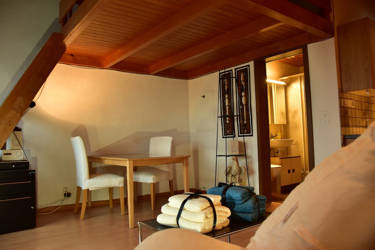 A cozy studio layout features a small dining table with two white chairs and a kitchenette. Towels are neatly arranged on a coffee table. A staircase leads to a sleeping area above, while a bathroom doorway is visible in the background.