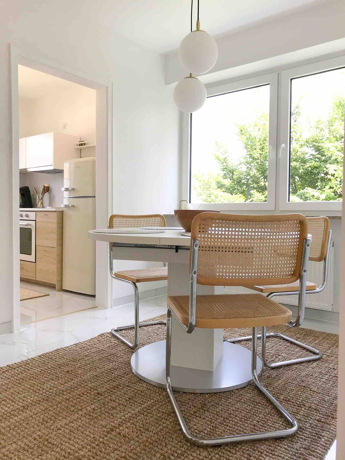 A bright dining area features a round table surrounded by three woven chairs. Natural light floods through large windows, illuminating the simple decor. A woven rug is placed underneath, adding a cozy touch to the space, while a glimpse of a modern kitchen is seen in the background.