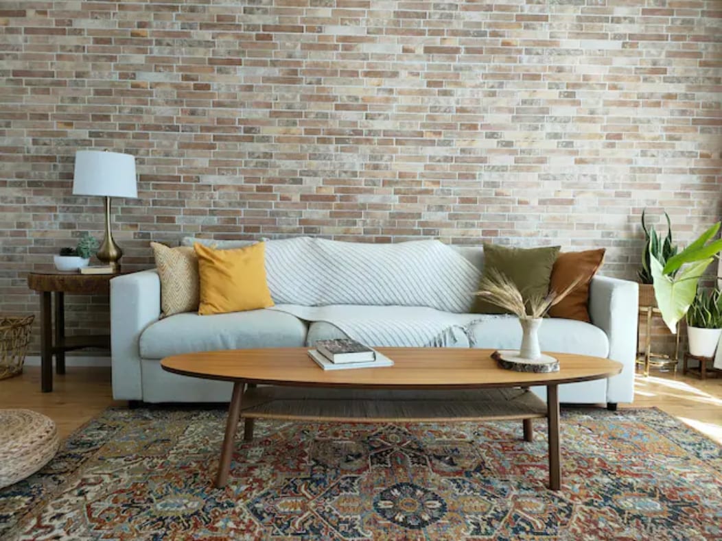 A light-colored sofa with decorative cushions sits in front of a textured brick wall. A wooden coffee table is positioned in front of the sofa, complemented by a patterned area rug. A lamp stands beside the sofa, and a plant is visible in the corner.