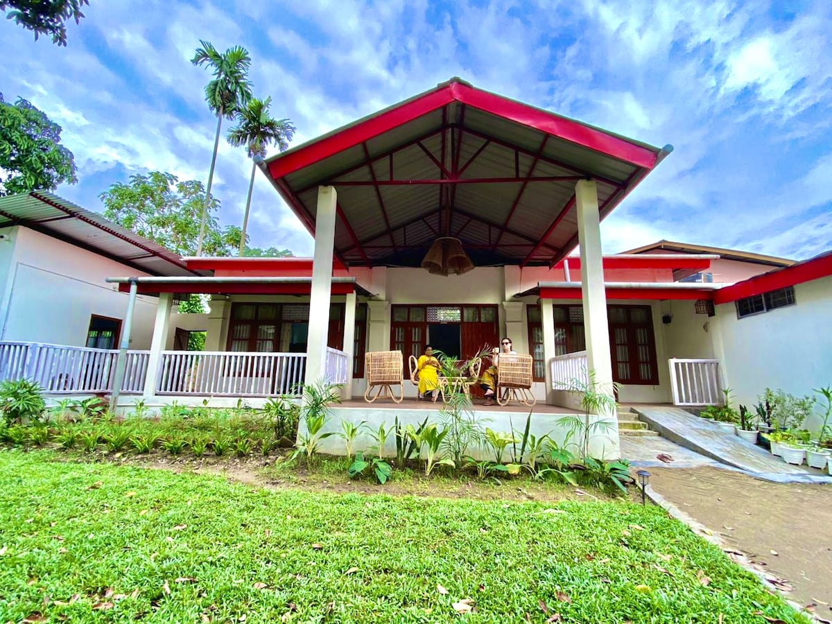 The traditional Assamese-style villa features a spacious veranda with seating arrangements, surrounded by a well-maintained lawn. Red-roofed architecture contrasts with the greenery, enhancing the welcoming entrance. The structure is framed by tall palm trees under a partly cloudy sky.