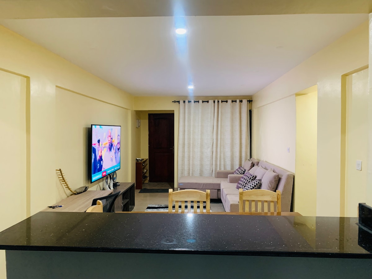 A spacious living room features comfortable furniture, including a sofa and chairs. A large smart flat-screen TV stands on a low cabinet. Light-colored curtains frame a window, enhancing the bright ambiance. A dark countertop is visible in the foreground, creating a clear division between spaces.