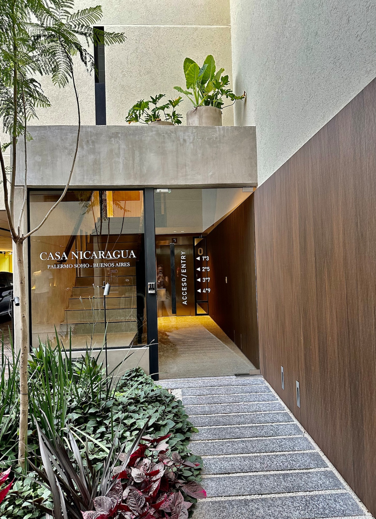 The entrance to Casa Nicaragua is depicted, featuring a sleek glass door framed by wooden accents. Lush greenery and decorative plants are positioned on the balcony above, while stone steps lead to the entrance, surrounded by vibrant foliage and modern architecture.