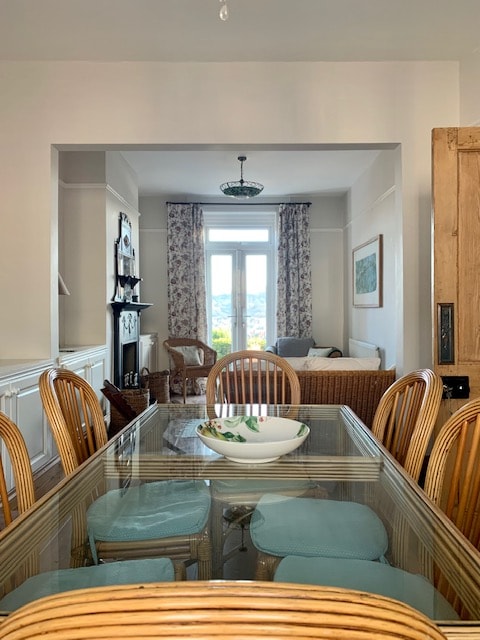 A dining area is seen featuring a long table surrounded by rattan chairs. Natural light streams through a large window at the far end, illuminating the room. A glass bowl is positioned at the center of the table, while floral curtains frame the view beyond.