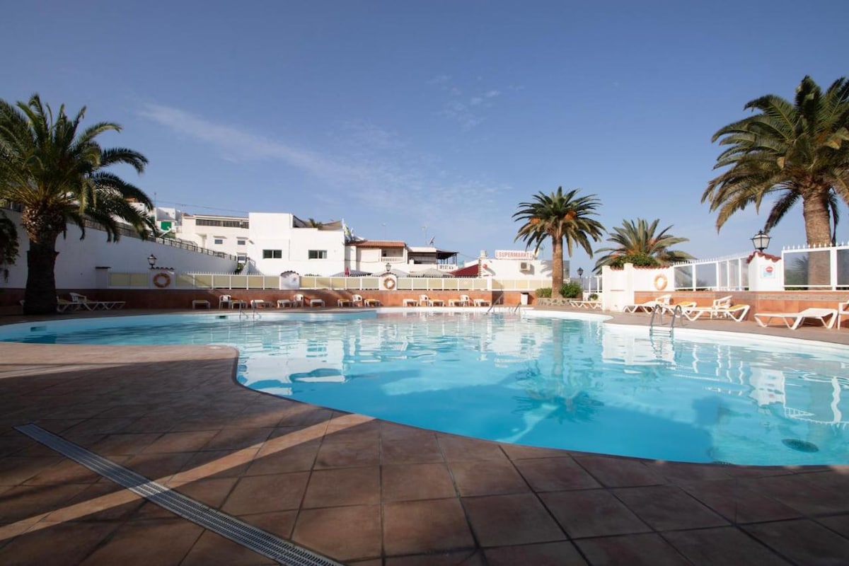 A clear swimming pool is surrounded by palm trees and sun loungers. The area is well-lit, with white buildings visible in the background, providing a serene setting for relaxation. The blue water reflects the bright sky, enhancing the tranquil atmosphere.