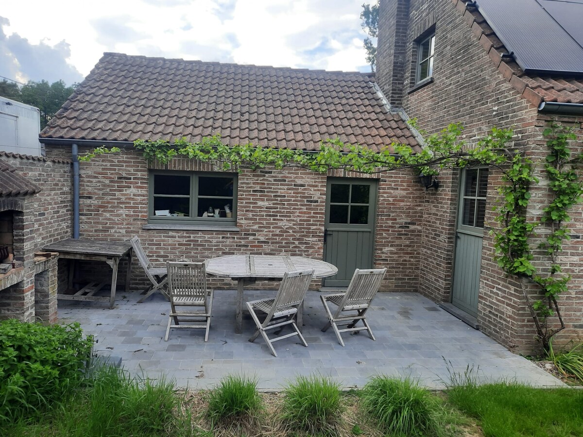 An outdoor terrace features a large round table surrounded by several wooden chairs. Climbing plants adorn the brick wall, while a stone pathway leads to the lush green garden, providing a peaceful space for gatherings.