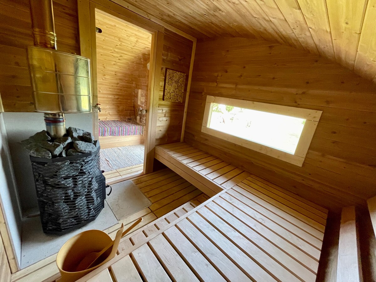 A wooden sauna interior features a stone fireplace and wooden benches arranged for relaxation. A window allows natural light to enter, illuminating the warm wooden walls and enhancing the serene environment.