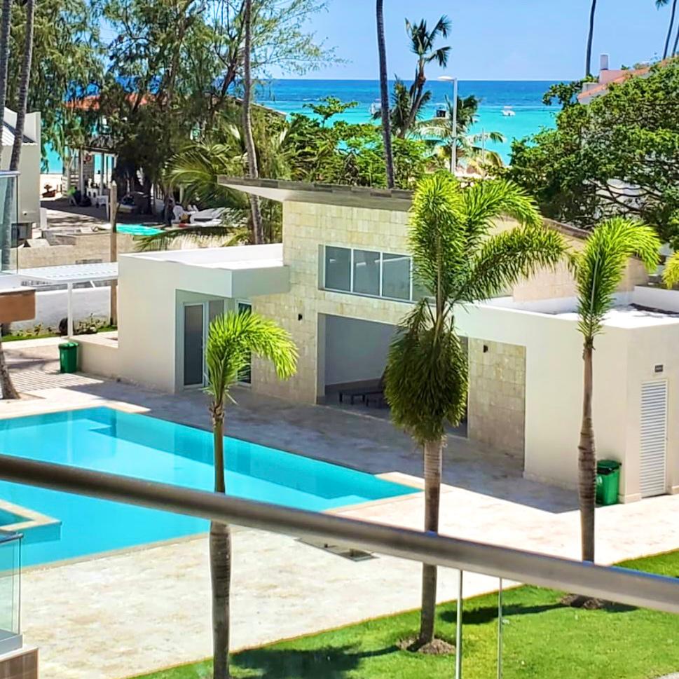 A clear view of a serene swimming pool is framed by lush palm trees. The pool reflects the bright blue sky, while distant ocean waves can be seen beyond a modern building with large glass windows. Well-maintained greenery surrounds the area.