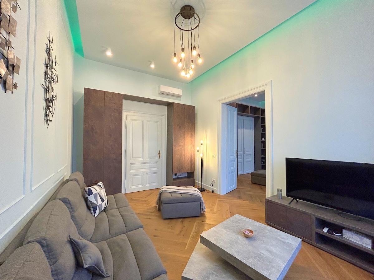 A modern living area features a comfortable sectional sofa and a coffee table. A large television is placed on a low console. Soft lighting is provided by a decorative chandelier and ambient LED lighting along the ceiling. Two doors lead to additional spaces.