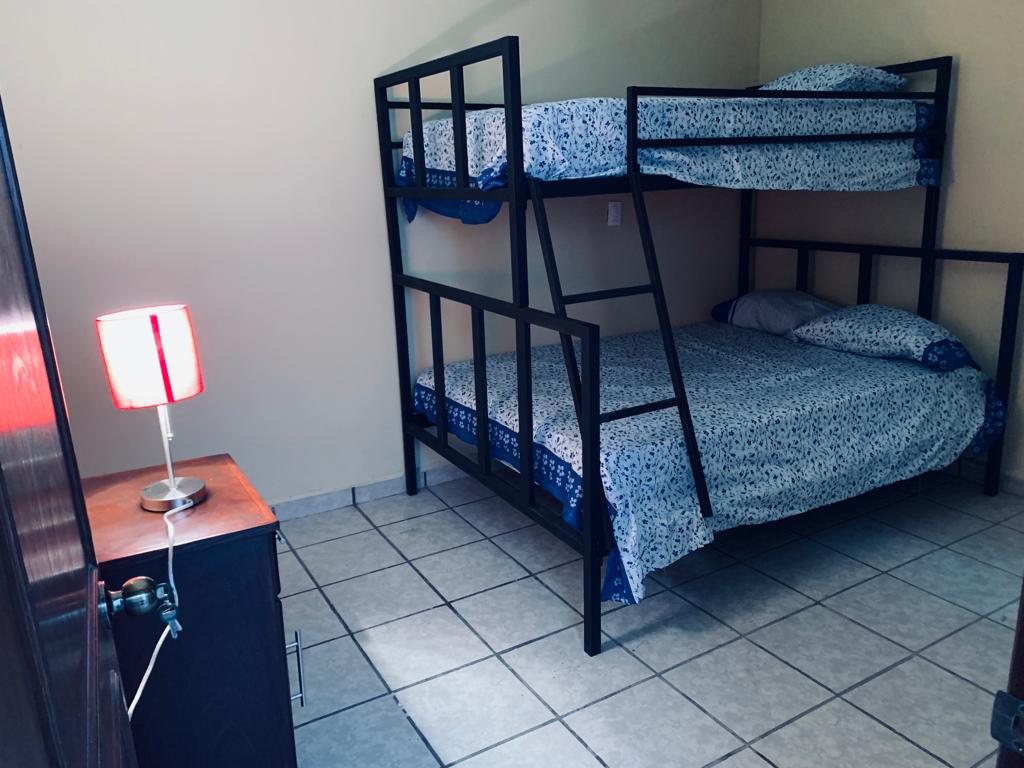 A simple bedroom features a metal bunk bed with patterned bedding. A spacious lower bed is complemented by a small nightstand. A bedside lamp with a fabric shade provides soft lighting, while tiled flooring adds a clean, functional touch to the room.
