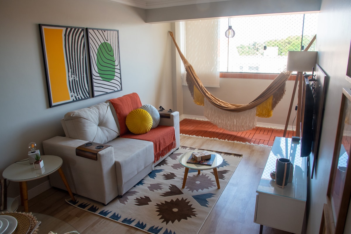 A welcoming living area features a comfortable sofa adorned with colorful cushions and a nearby hammock. Artwork hangs on the walls, complemented by a patterned rug and a coffee table. Natural light fills the space through the window, creating a bright and airy atmosphere.