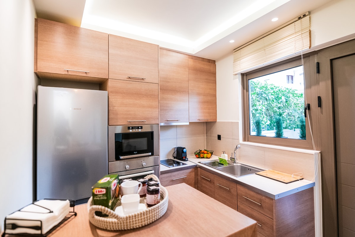 A modern kitchen is presented with light wood cabinetry and a sleek countertop. Appliances include a refrigerator, oven, and coffee maker. Natural light filters in through a window, providing a view of the outdoor area. Essential items for coffee preparation are neatly arranged on the counter.