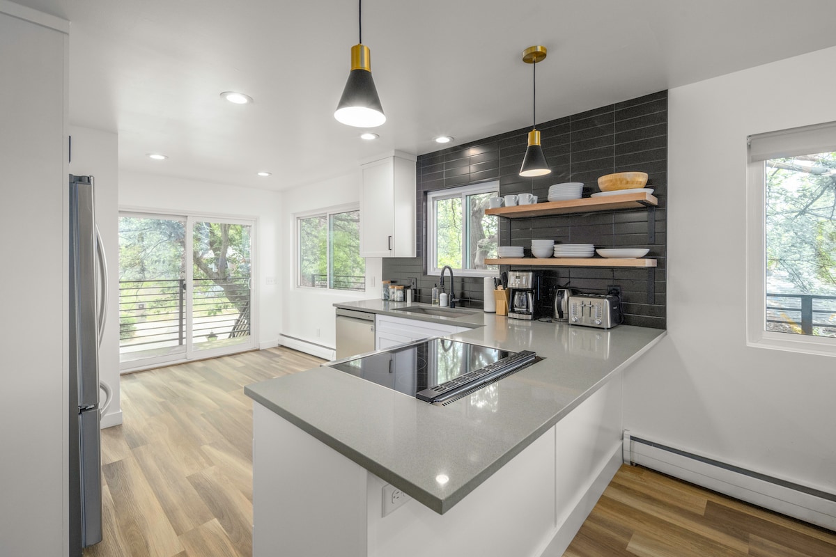 A modern kitchen features sleek cabinetry and a spacious countertop with a stovetop. Natural light pours in from large windows overlooking a deck, enhancing the bright and airy atmosphere. Open shelving displays dishware, and appliances include a coffee maker and toaster.