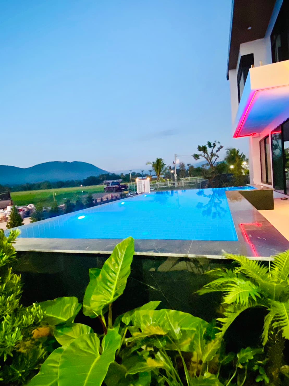 An infinity pool reflects the evening sky, surrounded by lush greenery and palm trees. Soft lighting enhances the tranquil vibe, while distant mountains create a scenic backdrop. The modern architecture of the building complements the outdoor space, inviting relaxation and enjoyment.