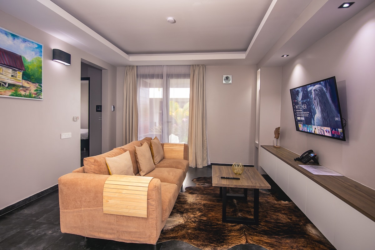 A spacious living room features a comfortable sofa and a wooden coffee table. Soft lighting highlights the soothing color palette, while a smart TV is mounted on the wall, providing entertainment options. Large curtains frame a window, allowing natural light to fill the room.