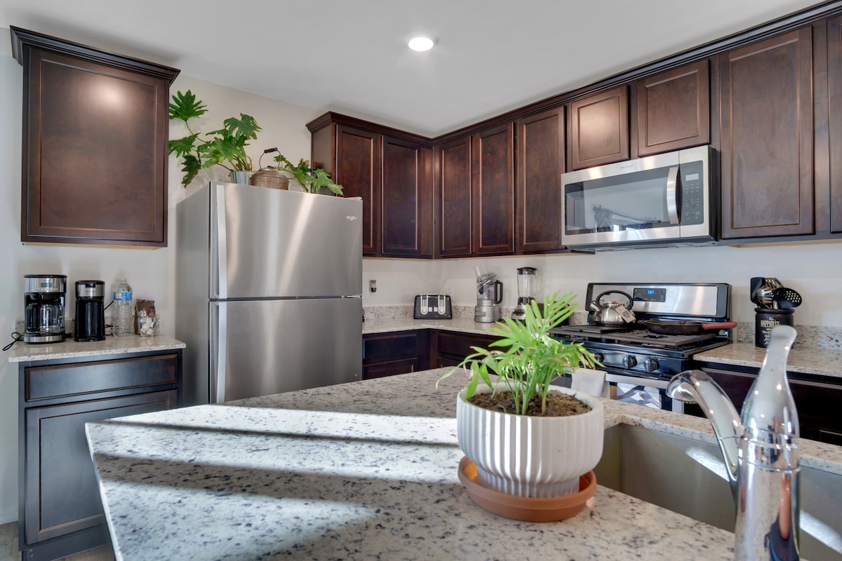 A modern kitchen is featured with dark wood cabinetry and stainless steel appliances. A granite countertop is complemented by a small potted plant, while a coffee maker and toaster are visible. Bright lighting enhances the space, creating a functional cooking area.