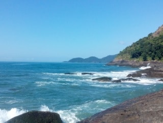 The image captures a tranquil coastal view featuring rolling waves gently crashing against rocky outcrops. A distant mountain range is visible under a clear blue sky, enhancing the serene atmosphere of the beachfront location.