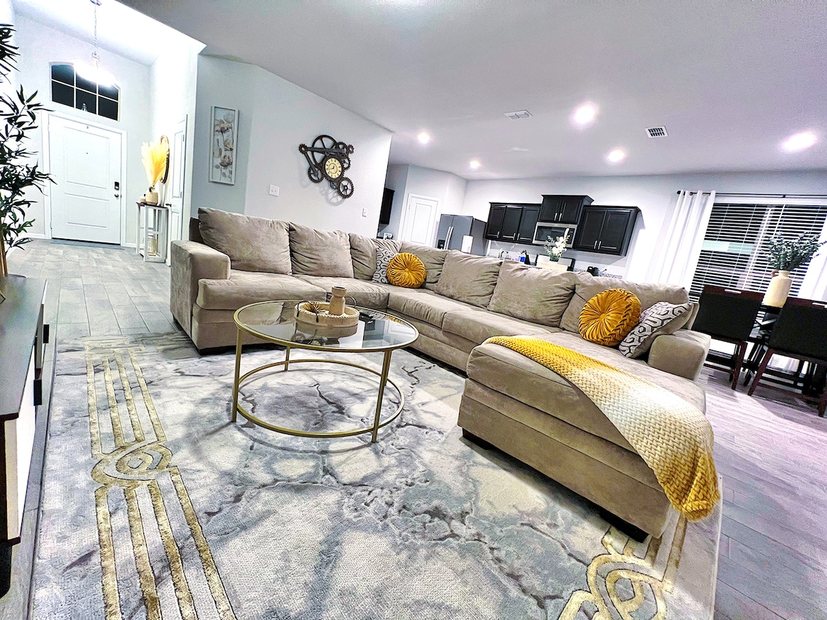 A spacious living room is presented with a large sectional sofa featuring decorative cushions. A round coffee table sits at the center, complemented by a patterned area rug. Light from nearby windows brightens the area, while a dining table is visible in the background.