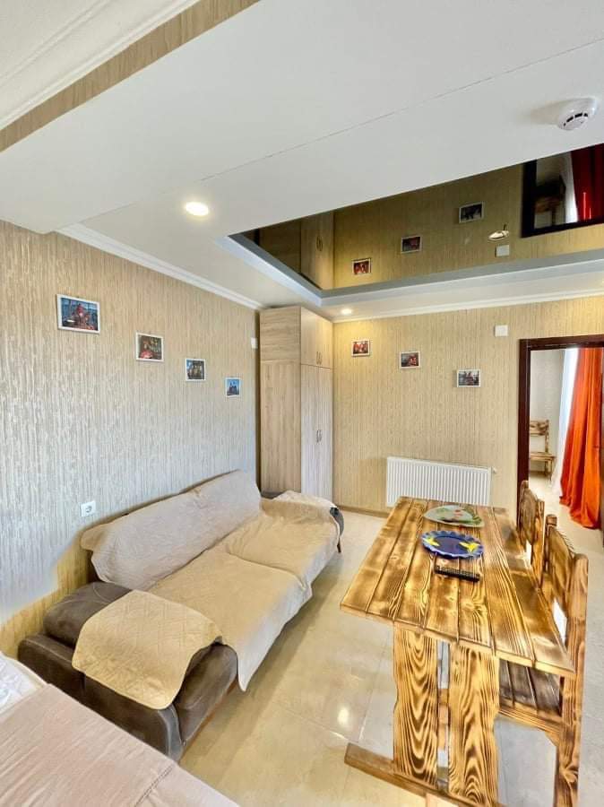 A living area is featured with soft furnishings, including a light-colored sofa and a wooden dining table. The table is set for dining, and framed photographs adorn the wall. Bright orange curtains frame a window, enhancing the room's warm ambiance.