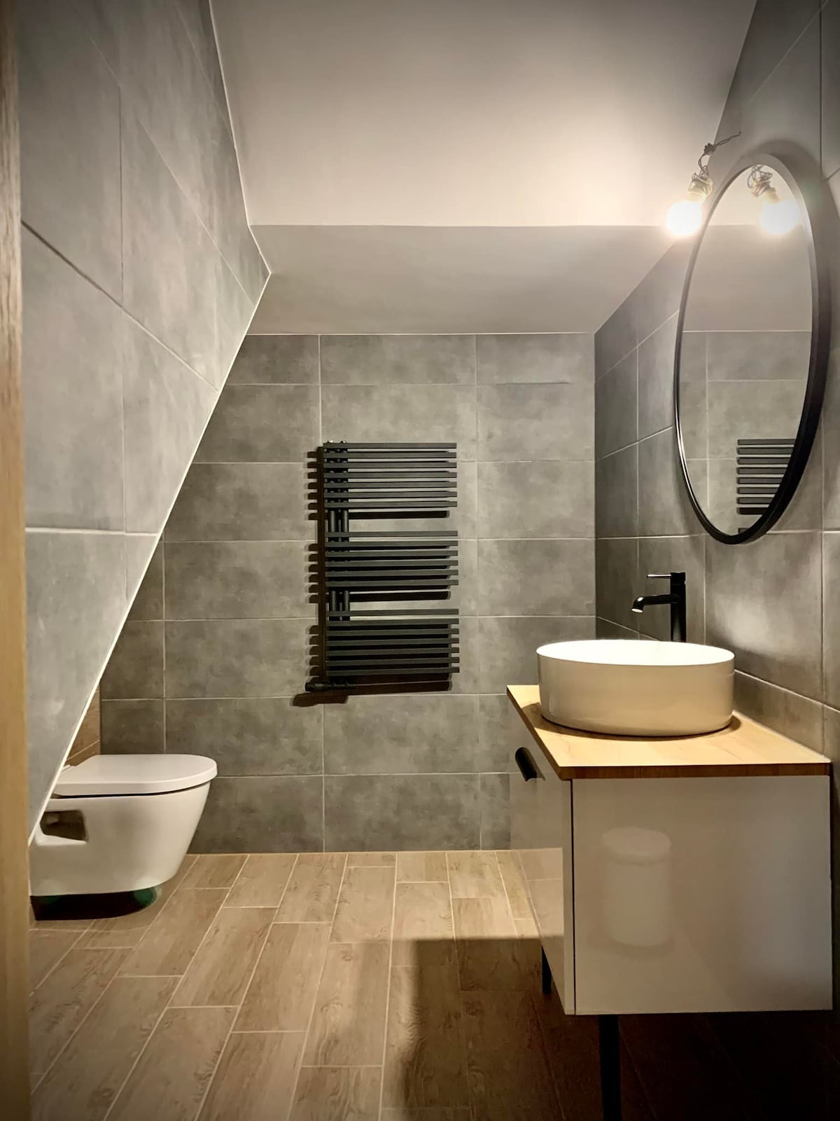 A modern bathroom features sleek gray tiles covering the walls and floor. A circular mirror hangs above a minimalist sink on a wooden vanity. A towel radiator is mounted on the wall, and a toilet is positioned to the side, creating a functional and organized space.