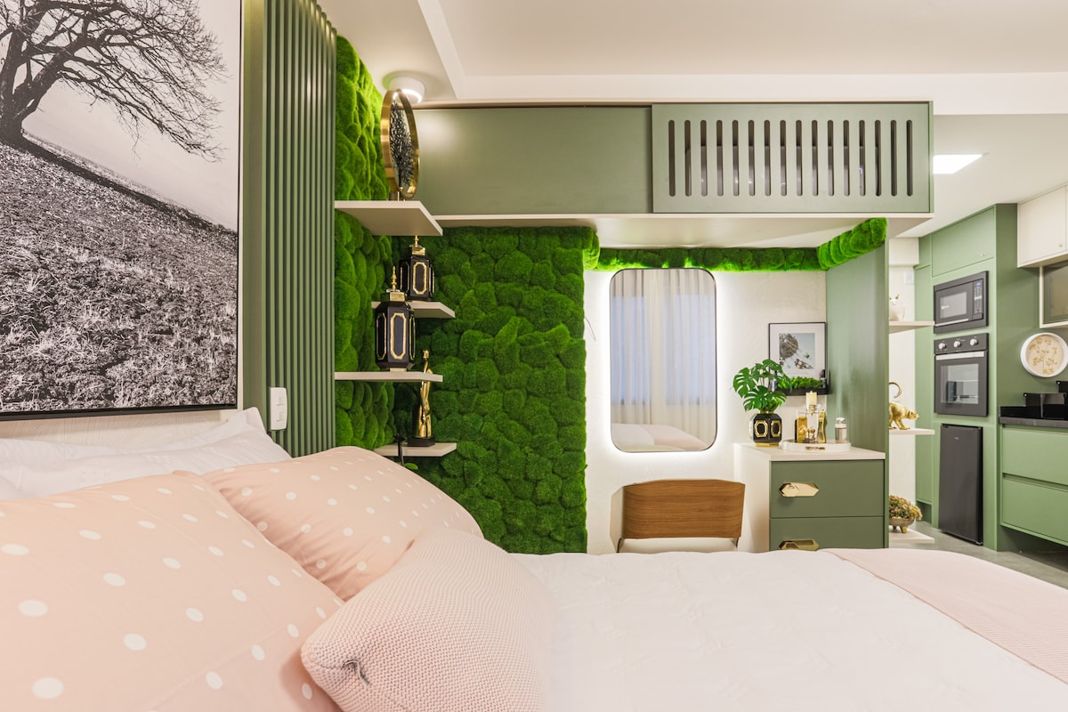 A serene sleeping area is highlighted by soft pink bedding with white polka dots. An accent wall of vibrant green moss complements a modern design and enhances the warmth of the space. Natural light filters in through a round window, softly illuminating the room.