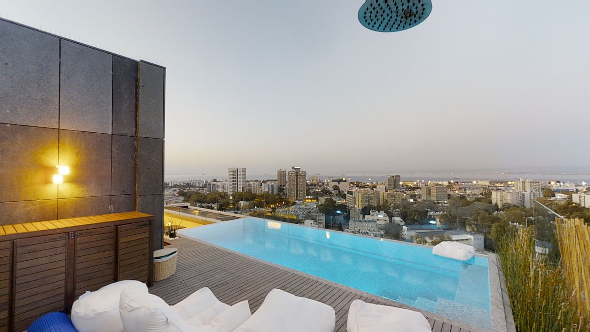 An infinity pool offers stunning views of the city skyline at dusk. Light from a nearby lamp casts a warm glow over a seating area with plush cushions. The horizon displays a gradual transition from blue to soft orange, creating a serene backdrop.