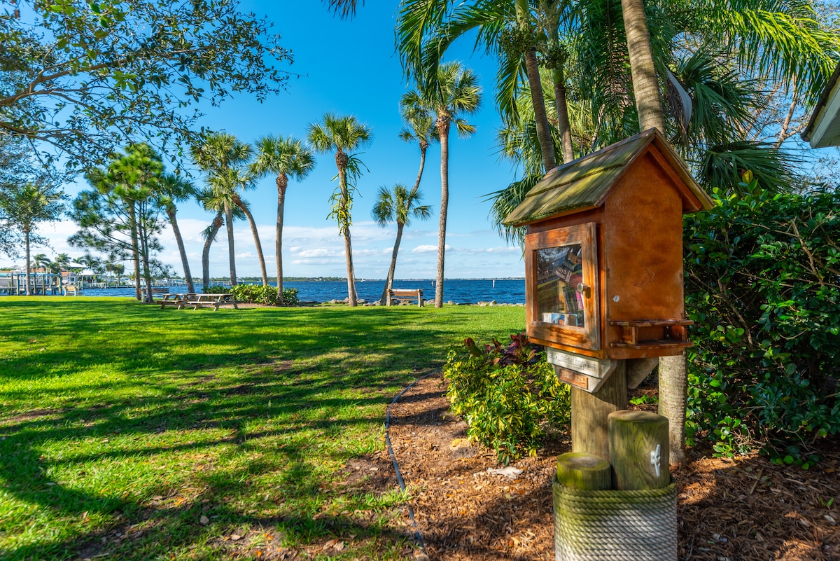 A scenic riverside park is depicted, with a view of the water through swaying palm trees. A small wooden kiosk stands on a grassy area, surrounded by lush landscaping. Benches are positioned nearby, offering a place to relax and enjoy the natural surroundings.