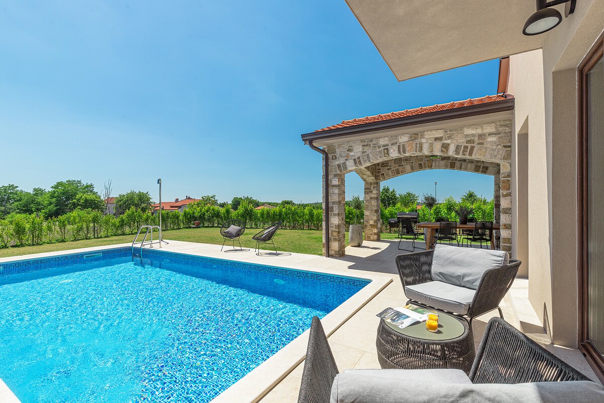 A modern outdoor area includes a well-maintained swimming pool, framed by a stone-finished terrace. Lounge chairs are positioned nearby, facing the water, while a dining area is shaded under a pergola. Lush greenery surrounds the space, creating a serene environment.