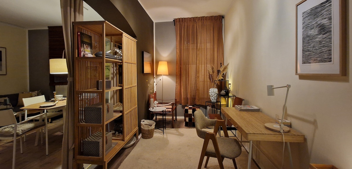 A shared living area features a wooden bookshelf filled with various items and a small desk. Soft lighting from a lamp enhances the space, while a light-colored rug covers the floor. A cozy seating area with a small table is visible in the background.