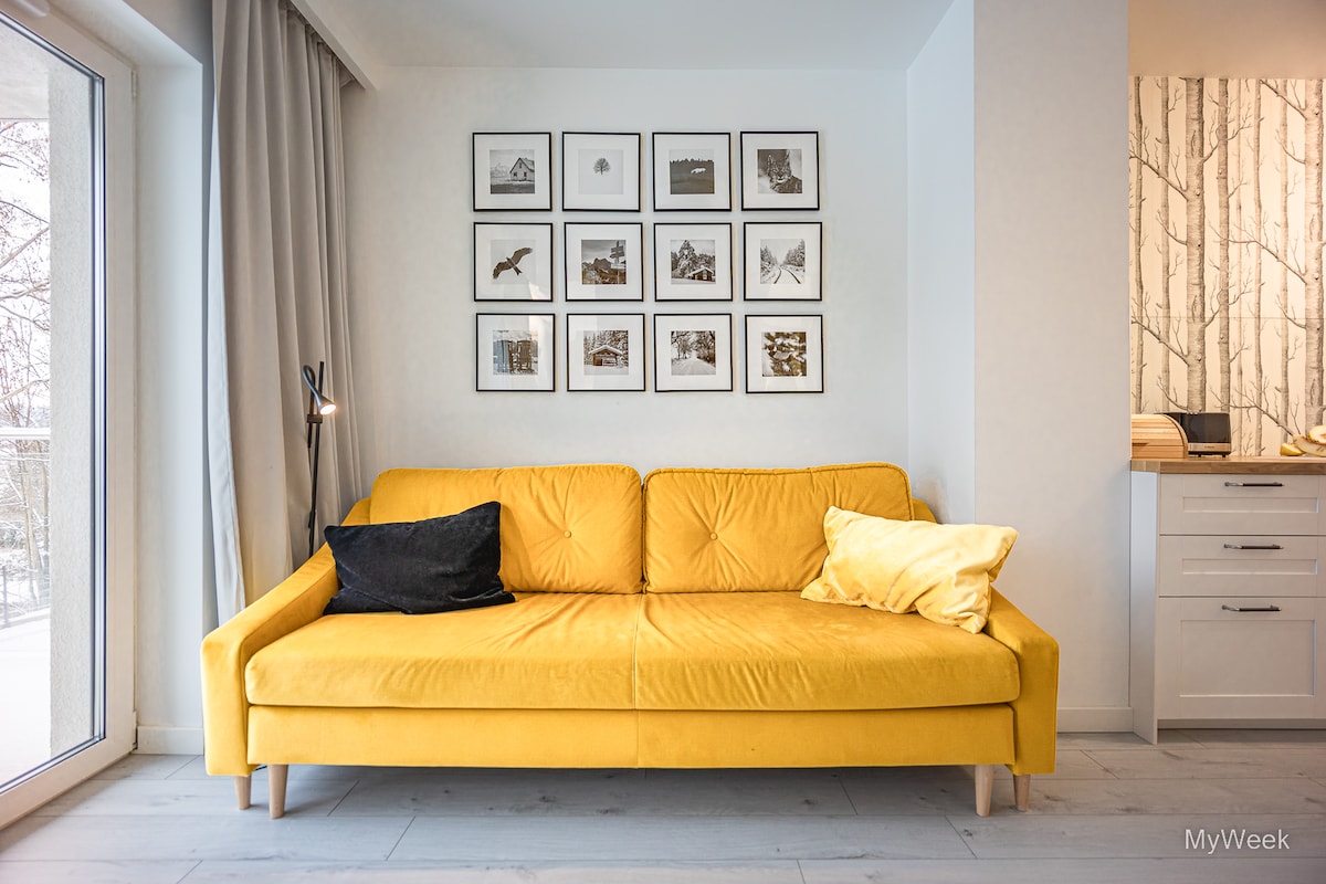 A bright yellow sofa with a black cushion is positioned against a light-colored wall adorned with a grid of framed black and white photographs. Natural light is filtered through a large window draped with soft gray curtains.