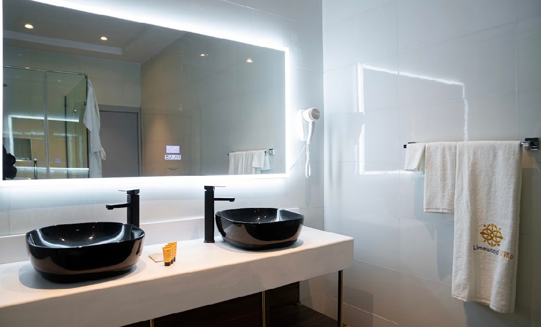 An elegant bathroom is equipped with a double vanity featuring two black vessel sinks. A large illuminated mirror hangs above, reflecting the modern fixtures. Fresh white towels are neatly displayed on a towel rack, contributing to a clean and spacious environment.