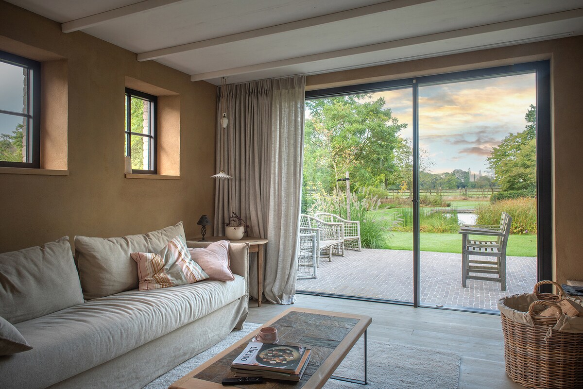 A lounge area features a spacious sofa adorned with soft cushions, with a wooden coffee table in front. Large sliding glass doors open to a serene garden view, showcasing a patio with outdoor seating and lush greenery surrounding a swimming pond.