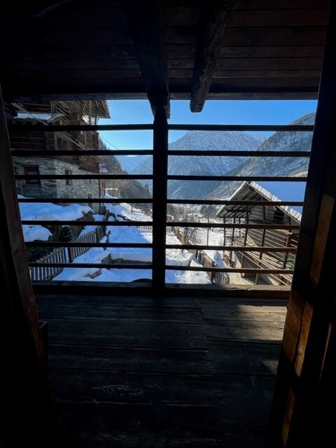 A wooden balcony presents a view of snow-covered mountains and rustic buildings below. The railing consists of horizontal wooden slats, allowing for an unobstructed sightline into the tranquil landscape. Sunlight filters gently through the space, enhancing the serene atmosphere.
