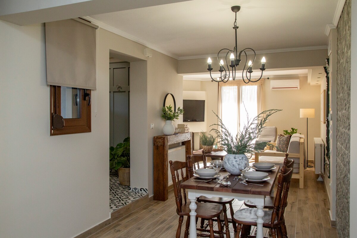A dining area is featured with a wooden table set for meals, surrounded by four matching chairs. Natural light enters through a nearby window, enhancing the spacious layout. A decorative chandelier hangs above the table, adding to the room's inviting ambiance.