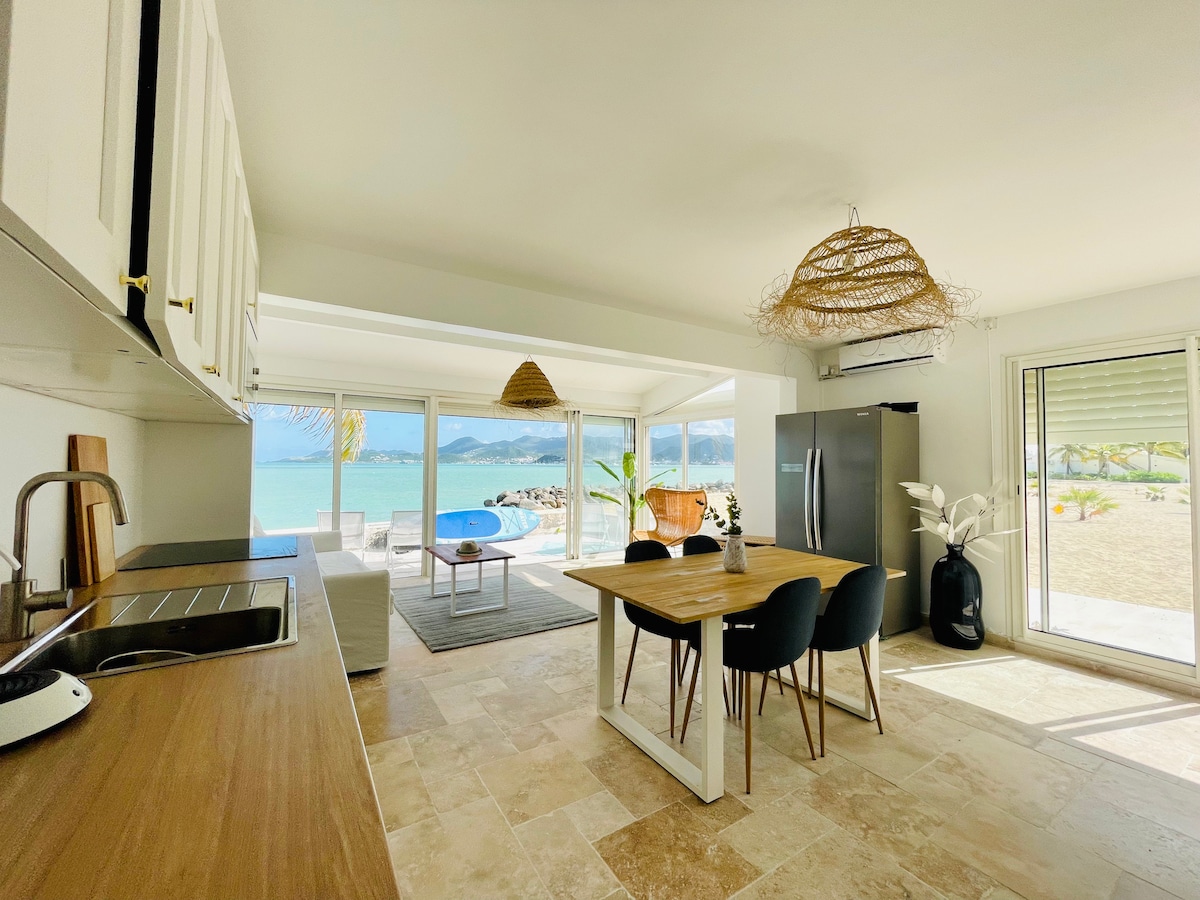 The modern kitchen features sleek cabinetry and a wooden countertop, complemented by a dining area with a table and black chairs. Large glass doors allow natural light to flood the space while offering views of the serene beach and ocean beyond.