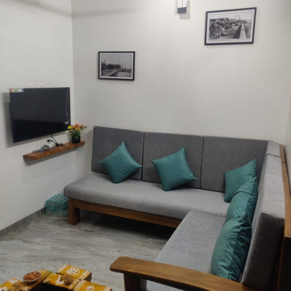 A compact living area is presented with a comfortable wooden sofa adorned with teal cushions. A wall-mounted television is situated above a wooden shelf, which holds a small vase of flowers. Monochrome photographs hang above, complementing the neutral color scheme.