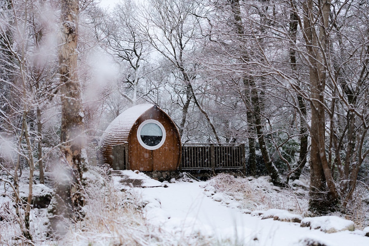 A wooden lodge with a circular window is nestled in a serene snowy landscape. Surrounding the lodge, trees are lightly dusted with white snow. A path leads to the entrance, and a deck extends from the side, offering a cozy outdoor space.