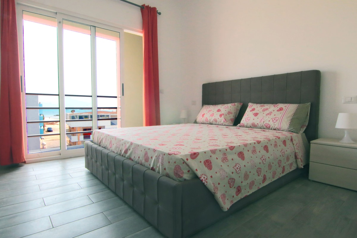 A modern bedroom features a spacious bed adorned with a playful pink heart pattern. Natural light fills the room through large sliding doors that open to a balcony, framed by soft coral curtains. A bedside table is positioned beside the bed, offering a cozy touch.