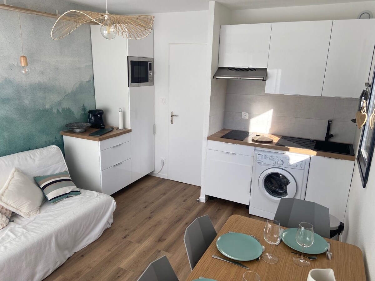 A modern kitchen area features sleek white cabinetry and a wood countertop. A small dining table with six chairs is set with glassware, while a comfortable sofa provides additional seating. Natural light illuminates the space through an adjacent window, creating a welcoming atmosphere.