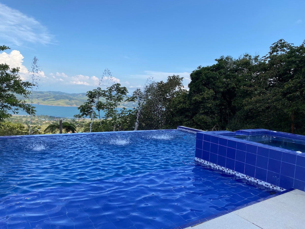 An infinity pool is visible with shimmering blue water, offering a view of the expansive landscape that includes rolling hills and a lake in the distance. Surrounding foliage provides a natural backdrop, enhancing the sense of tranquility and connection to nature.