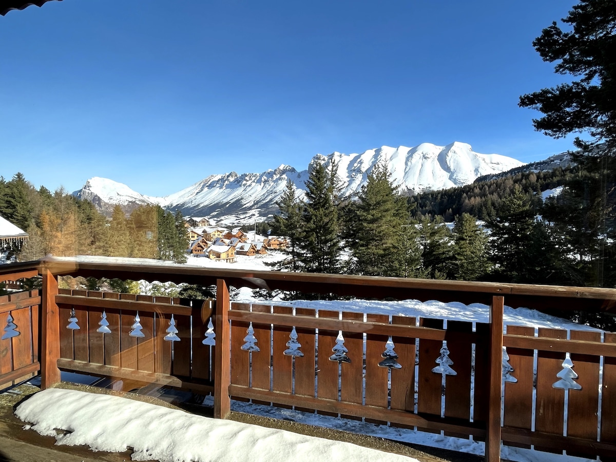 A wooden balcony railing is adorned with festive decorations, overlooking a snow-covered landscape. Majestic mountains rise in the background, while a charming village is nestled among the trees below, under a clear blue sky.