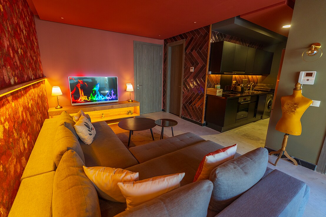 A spacious living area is displayed, featuring a comfortable sectional sofa adorned with decorative pillows. A vibrant TV with animated colors is positioned on a wooden console. The sleek kitchen area is partially visible, showcasing modern cabinetry and a herringbone tiled wall.