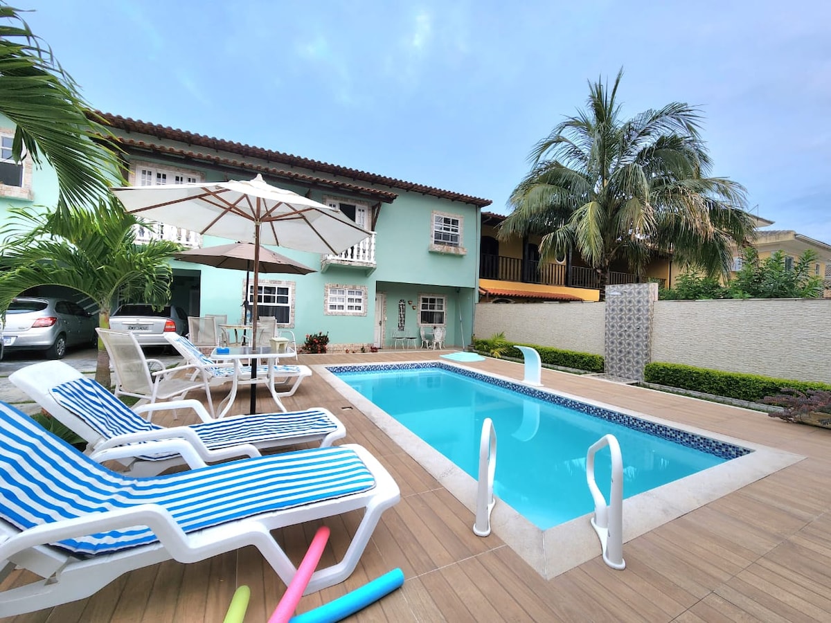 A serene outdoor area features a clear swimming pool surrounded by wooden decking. Lounge chairs with striped cushions are arranged around the pool, complemented by a large umbrella. Tropical plants line the space, enhancing the inviting atmosphere of the garden.