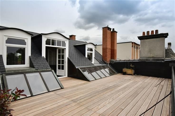A private rooftop terrace features wooden decking and several skylights allowing natural light to enter the space. The area is framed by slanted roofs and showcases a view of nearby chimneys and buildings, creating a sense of openness.
