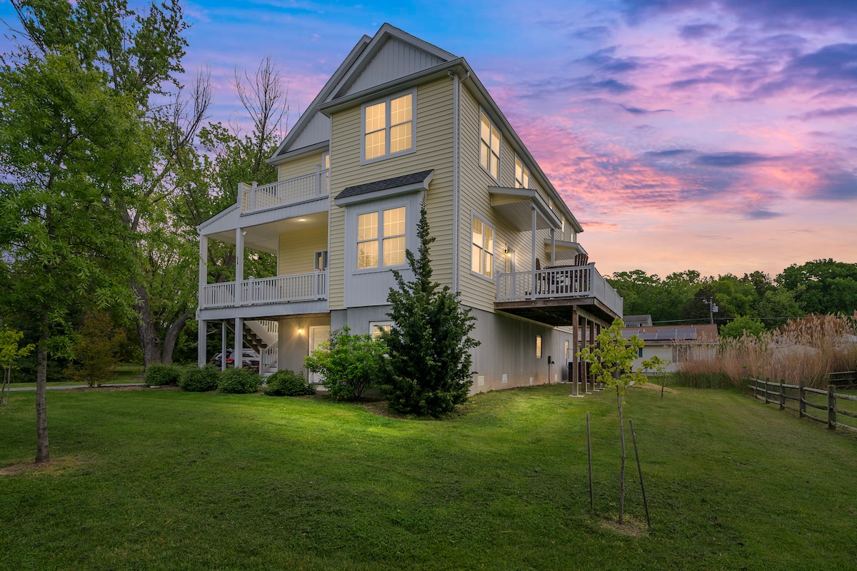 The exterior of a spacious, two-story home is highlighted by soft exterior lighting at dusk. A wraparound porch is visible, surrounded by neatly landscaped greenery. The tranquil setting is enhanced by a sky painted in hues of pink and purple, creating a serene atmosphere.