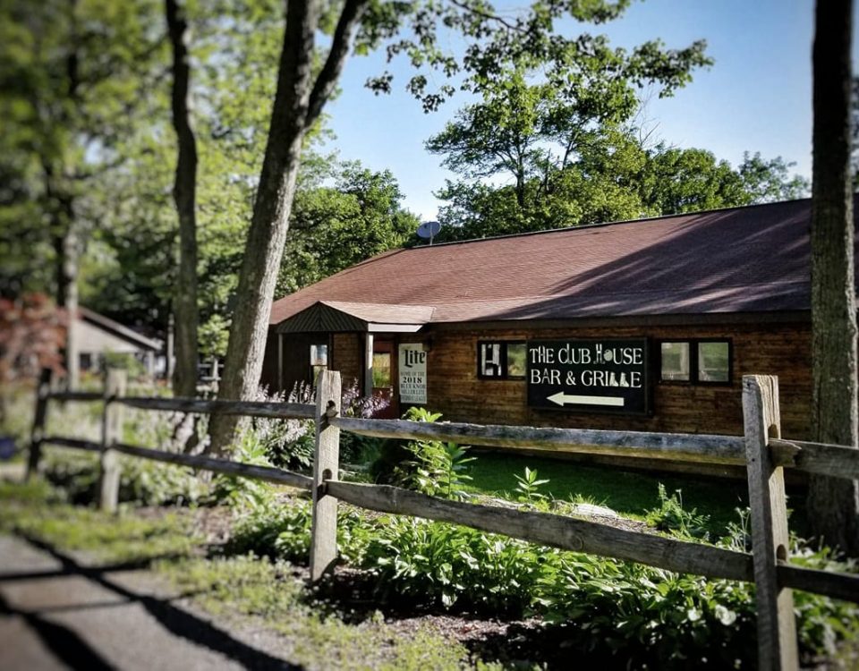 The Clubhouse Bar and Grill is set amidst greenery, featuring wooden siding and a traditional roof. A fenced entrance provides a rustic charm, while surrounding trees add to the natural ambiance. Signage is prominently displayed, indicating the venue's name and offerings.
