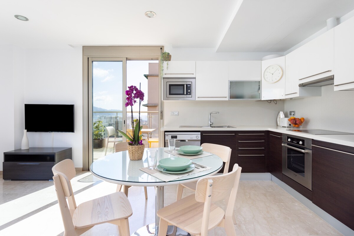 A modern kitchen area is illuminated by natural light streaming through a large window. A circular glass dining table is set for two, accompanied by four light wooden chairs. White cabinets and stainless steel appliances are visible, along with a television mounted on the wall.