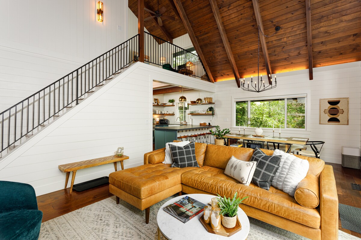 An inviting great room features a spacious, tan leather sectional sofa with various cushions arranged on it. A round coffee table displays greenery and magazines. High ceilings with exposed wood beams complement the warm wood tones throughout, while a staircase leads to the upper level.