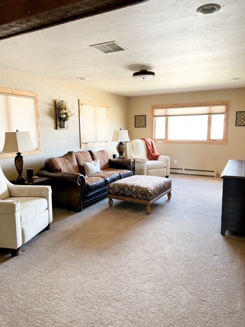 A spacious living area features comfortable seating arrangements, including two light-colored armchairs and a dark leather couch. A decorative ottoman sits in the center, and natural light filters through large windows dressed with wooden frames.